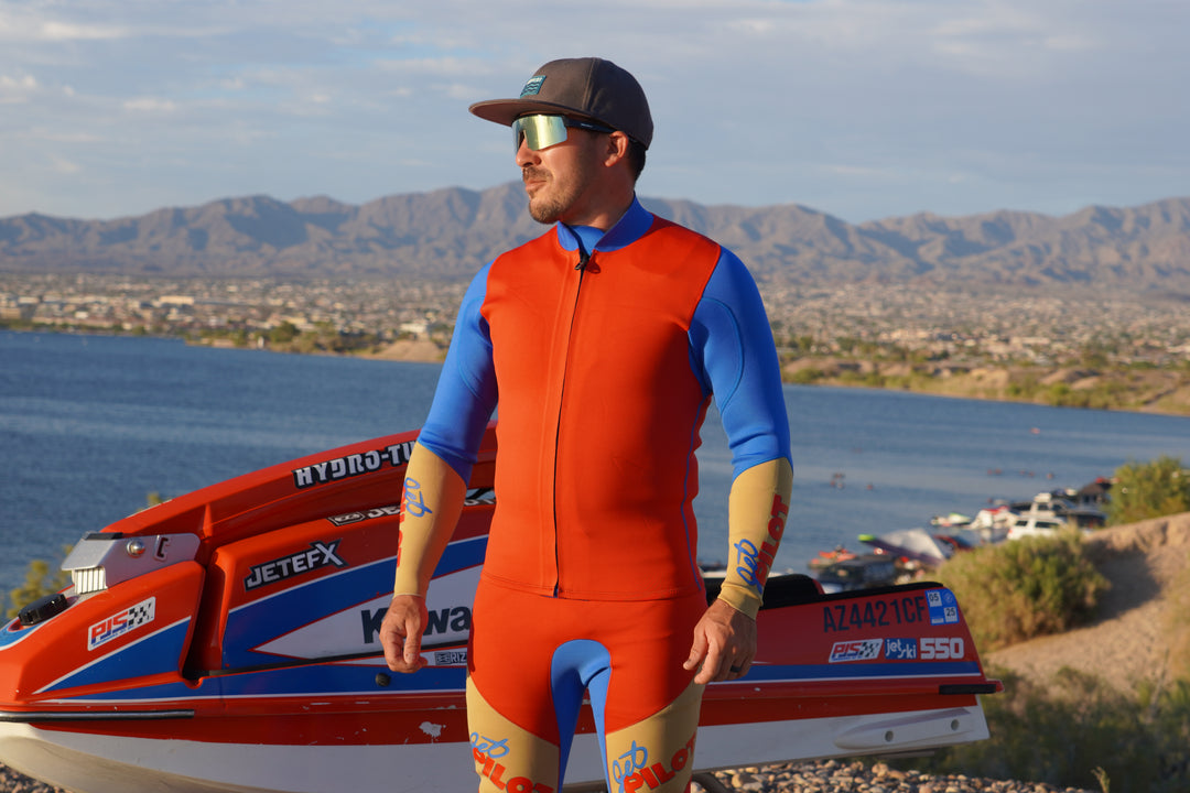 Mark Gomez in a red and blue Jetpilot Vintage Class wetsuit standing next to a jet ski with a scenic background. #color_red-blue