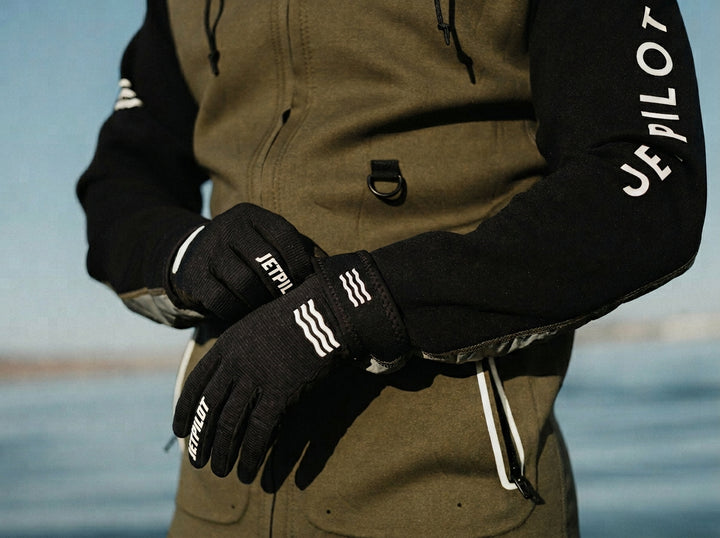 Person wearing black gloves and a brown jacket with 'JETPILOT' branding, standing by water. #color_black