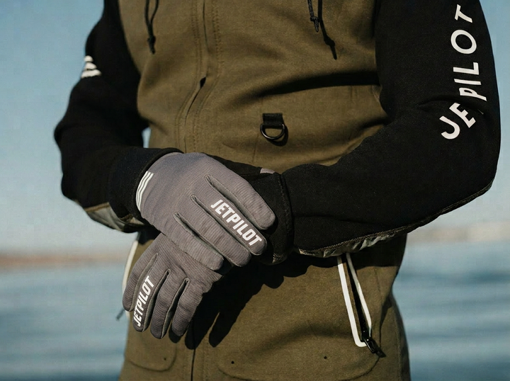 Person wearing a green jacket and gray gloves with 'JETPILOT' branding, standing by water. #color_charcoal