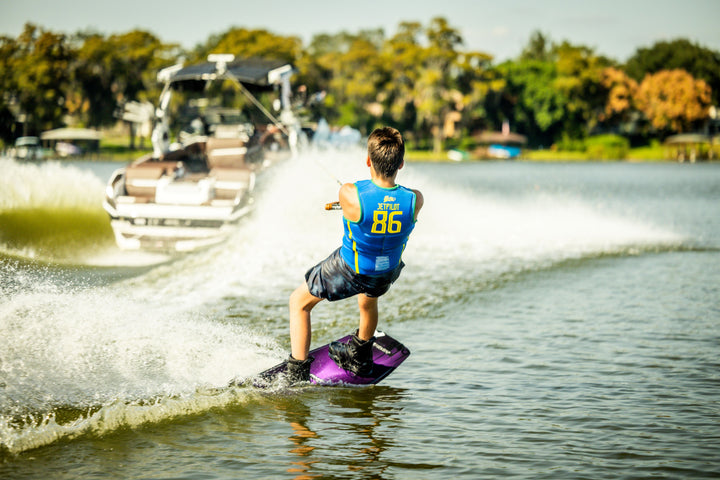 Person wakeboarding behind a boat on a lake with trees in the background. #color_yelbrz