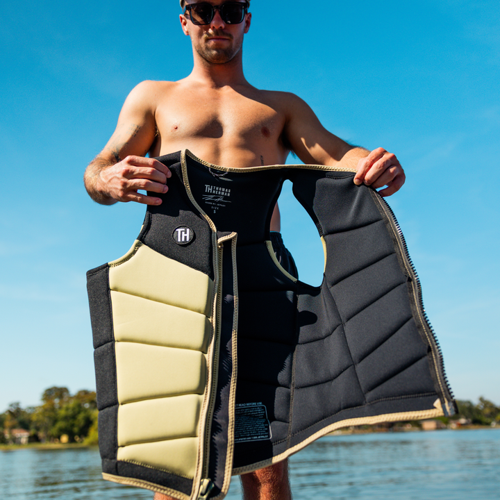 Man holding a black and beige life jacket by a body of water. #color_khaki