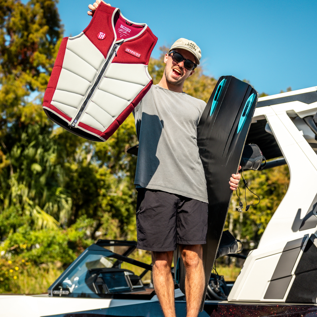 Man holding a life vest and a water board with a boat and trees in the background. #color_burgundy