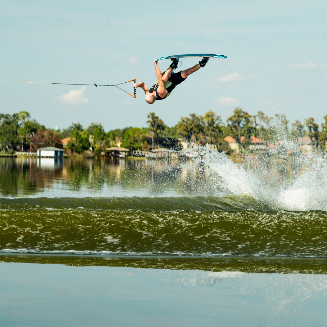 Person wakeboarding in mid-air over a lake with trees and houses in the background.  #color_khaki