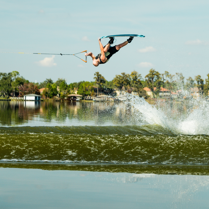 Person wakeboarding in mid-air over a lake with trees and houses in the background.  #color_khaki